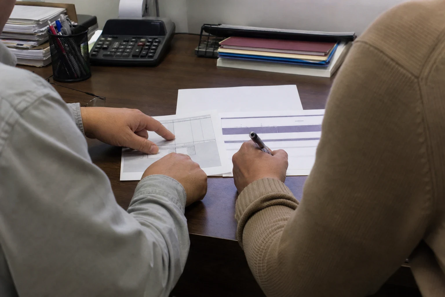 Two people seen from behind at a wooden office desk, one pointing at a printed form while the other holds a pen over a second document beside a calculator and folders